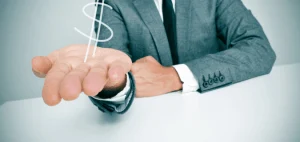 Person sitting at desk in suit with an outstretched hand that is holding dollar symbol.
