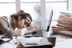 Woman sleeping at desk next to laptop and pile of files, glasses are askew on her face.