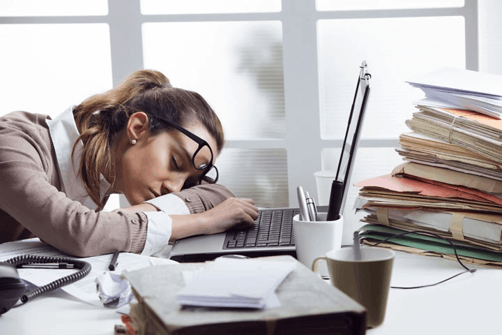 Woman sleeping at desk next to laptop and pile of files, glasses are askew on her face.
